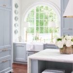 lauren-deloach-atlanta-showhouse-award-winning-kitchen-calacutta-marble-blue-painted-cabinets-brass-accents-farmhouse-sink-lamps-flanking-sink-hanging-plates